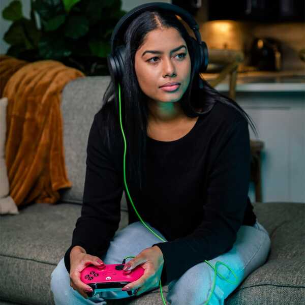 Young woman gaming with a pink Xbox controller and headphones in a cozy living room.