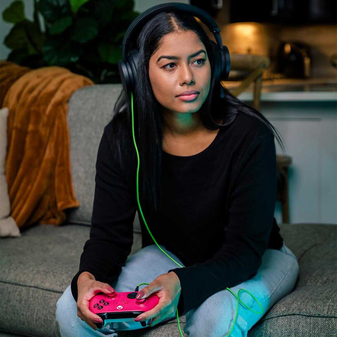 Young woman gaming with a pink Xbox controller and headphones in a cozy living room.