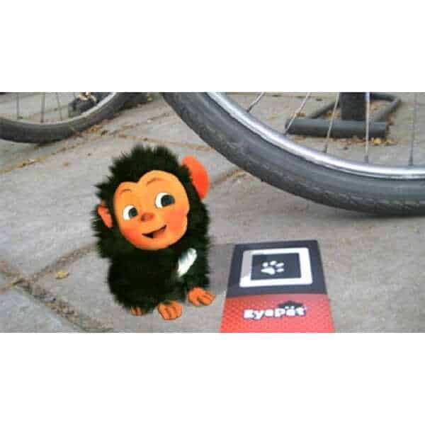 Cute virtual pet monkey from EyePet game on a sidewalk with a bike wheel nearby.