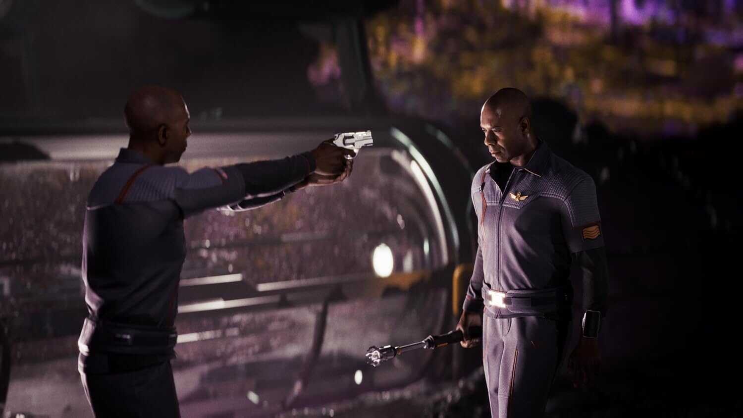 Two men in dark uniforms with a gun and a baton in a nighttime setting.