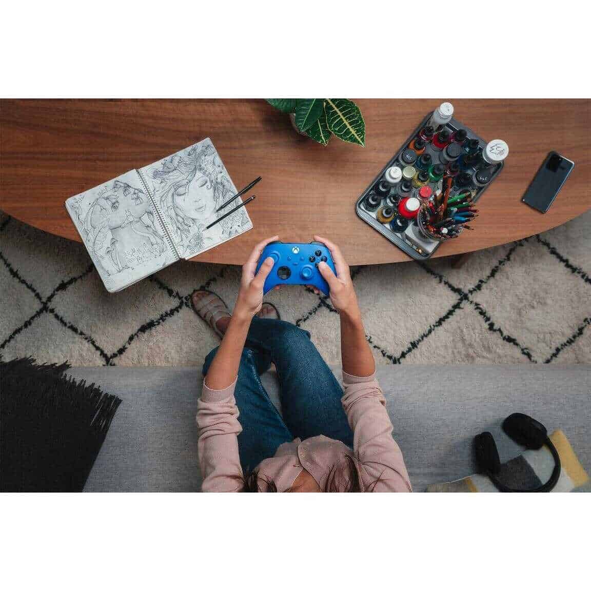 Person playing Xbox Series X controller with art supplies nearby.